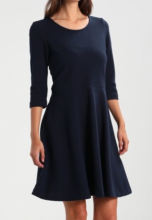 Femme portant une robe bleu marine moulante à texture avec des manches trois-quarts et une jupe évasée sur un fond uni.