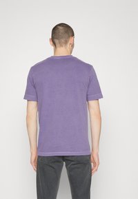 Fioletowa bawełniana koszulka z krótkim rękawem, okrągłym dekoltem i widocznymi detalami szwów. Nosi się z ciemnoszarymi dżinsami, widok z tyłu.