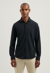 Hombre con una camisa de botones de manga larga oscura y pantalones claros beige, de pie contra un fondo claro y liso.