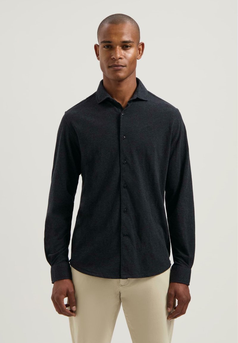 Hombre con una camisa de botones de manga larga oscura y pantalones claros beige, de pie contra un fondo claro y liso.