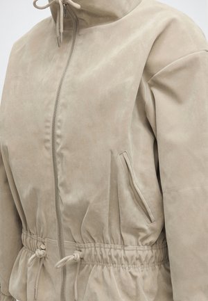 Veste en daim beige avec col montant, fermeture éclair devant, poches latérales et taille ajustable avec cordon.