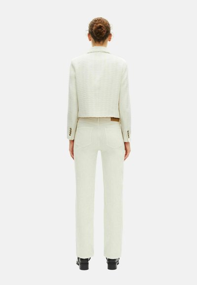 Femme aux cheveux bouclés en chignon portant une veste blanche texturée, un pantalon crème taille haute et des chaussures noires, se tenant de dos.