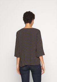 Kvinna med kort lockigt hår som bär stora örhängen, en marinblå topp med gula prickar och mörka jeans, sedd bakifrån.