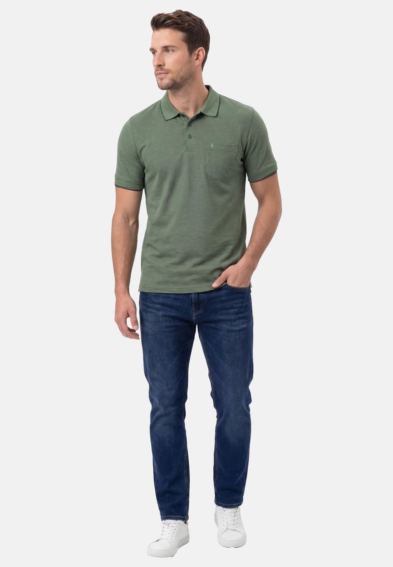 Uomo che indossa una polo verde a maniche corte, jeans blu scuro e scarpe da ginnastica bianche, in piedi con una mano in tasca, guarda verso sinistra.