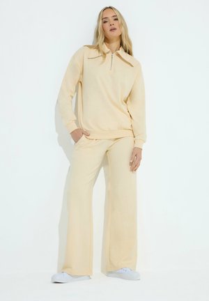 Vrouw met beige sweatvest met rits en wijde broek, gecombineerd met witte sneakers, staand tegen een effen lichte achtergrond.