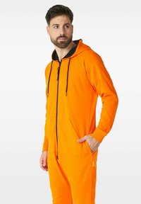 Oranžový zipový overal s černou kapucí, předními kapsami a stahovacími šňůrkami. Hladká textura a přiléhavý design s žebrovanými manžetami.