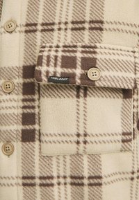 Beige and brown checkered flannel shirt with a soft texture, featuring a chest pocket and tan buttons. Brand label visible on the pocket.