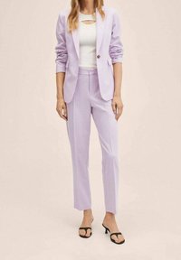 Completo in lilla composto da un blazer aderente con un solo bottone e pantaloni slim-fit, abbinato a una maglietta bianca con un design a taglio e sandali neri.