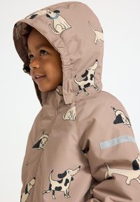 Veste marron clair avec des motifs de chiens, dotée d'une capuche, d'une texture lisse et d'une bande réfléchissante grise sur la manche.