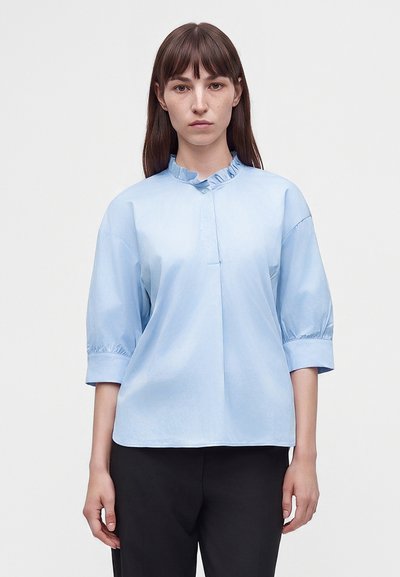 Femme aux cheveux bruns et raides porte un chemisier bleu clair à col volanté et manches trois-quarts ainsi qu'un pantalon noir, debout devant un fond uni.