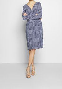 Abito blu marino con scollatura a V, maniche lunghe e un motivo geometrico; design al ginocchio. Indossato con sandali argentati a strappy.