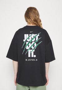 Svart oversized t-shirt med vit "JUST DO IT" slogan och gröna klospår. Tillverkad av bomull, med avslappnad passform och rundad hals.
