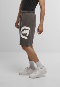 Gray shorts with an elastic waistband feature a large white graphic design on the left leg. White sneakers and ribbed socks complete the look.