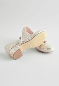 Glitzernde silberne Ballerinas mit runder Zehenpartie und kleinem Blockabsatz, ausgestattet mit einem Riemen und einer glatten, hellen Gummisohle.