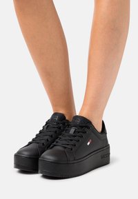 Persona che indossa sneaker in piattaforma nere con lacci e un piccolo logo rosso, bianco e blu sul lato, su uno sfondo neutro.