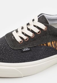 Baskets Vans noires avec lacets blancs, bout texturé, œillets marron, détail en grain de bois sur le côté, et semelle blanche sur fond blanc.