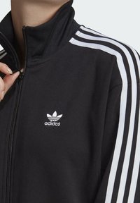 Černá bunda na zip z měkké látky, s bílými trojpruhovými akcenty na ramenou a malým bílým logem adidas na hrudi.