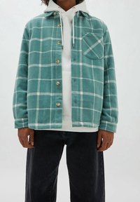 Modello che indossa una camicia a quadri turchese e bianca sopra un hoodie bianco, abbinata a jeans scuri, con uno sfondo neutro.