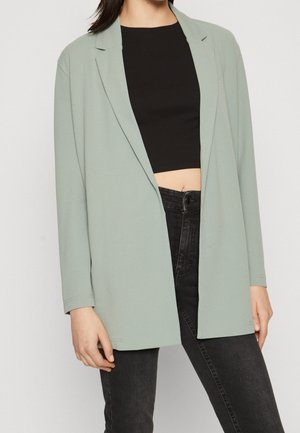Personne portant un blazer ouvert vert clair, un top court noir et un jean taille haute noir sur un fond uni.