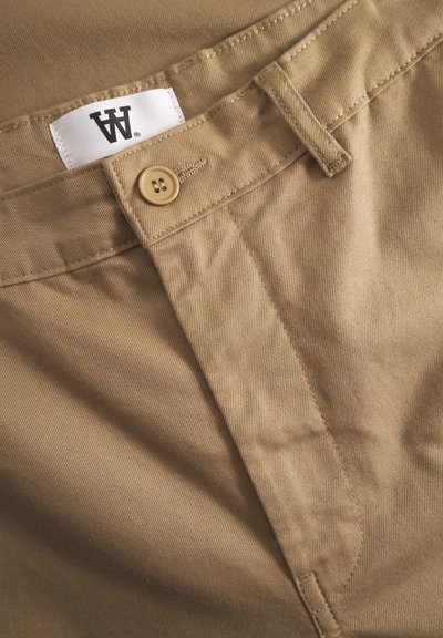 Tessuto di cotone color taupe con una texture liscia, dotato di un bottone e di un'etichetta cucita. Il design presenta una parte anteriore piatta e passanti per la cintura.
