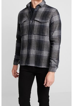 Homme portant une chemise à carreaux noire, grise et blanche sur un t-shirt noir avec un pantalon noir, debout devant un fond uni.