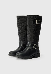 Botas de rodilla acolchadas en negro con un patrón texturizado, detalles de cuero, un cierre lateral de cremallera y una correa con hebilla en el tobillo.