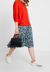 Czerwony sweter z dzianiny, kwiecista granatowa spódnica z białymi i zielonym akcentami, białe trampki z czerwonymi detalami oraz mała czarna torebka z drewnianym uchwytem.