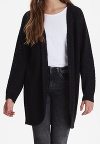 Černý pletený kardigan s otevřeným předním dílem, volným střihem, texturovaným vzorem a dlouhými rukávy, zkombinovaný s bílým tričkem a tmavými džíny.