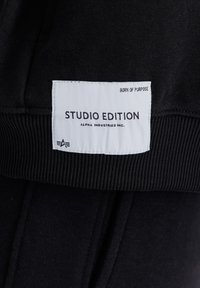 Crne trenirke s rebrastim pojasom i bijelom etiketom koja kaže "STUDIO EDITION" i "ALPHA INDUSTRIES INC."
