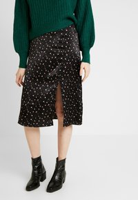 Černá květovaná sukně s předním rozparkem, vyrobená z hladké látky. Kombinována s černými kotníkovými botami s texturovaným povrchem. Navrchu zelený pletený svetr.