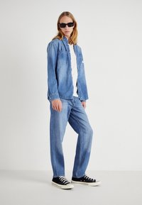 Niebieska koszula jeansowa w stylu zapinanym na guziki, z dwiema przednimi kieszeniami, zestawiona z luźnymi spodniami dżinsowymi w tym samym kolorze oraz czarnymi trampkami.