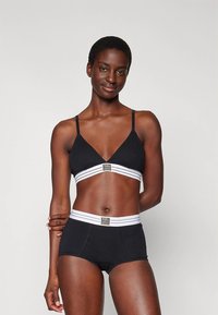 Bralette preta com alças finas e uma faixa larga com riscas brancas, combinada com calções pretos de cintura alta com uma faixa semelhante.
