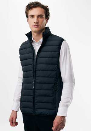 Ung mand iført en sort quiltet ærmeløs vest over en hvid skjorte med knapper mod en ensfarvet hvid baggrund.