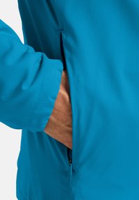 Giacca azzurro chiaro con superficie texturizzata e tasca a zip, caratterizzata da una vestibilità morbida e maniche lunghe. Gli accenti includono un tirazip visibile.
