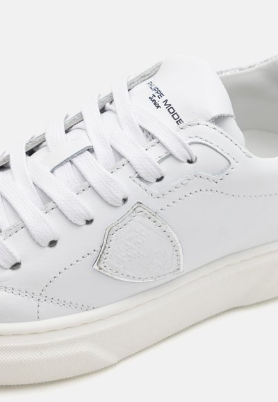 Baskets en cuir blanc avec des lacets plats blancs, semelles en caoutchouc et un accent métallique sur la languette. Présentent un logo en forme de bouclier sur le côté.
