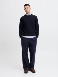 Marineblauer Strickpullover über einem hellblau-weißen gestreiften Hemd, kombiniert mit lockeren marineblauen Hosen und dunkelbraunen Schuhen. Einfaches, strukturiertes Design.