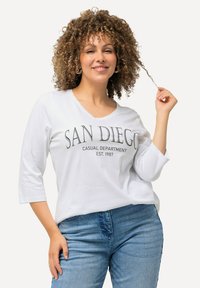 Weißes, locker sitzendes Hemd mit einem runden Ausschnitt, versehen mit der Aufschrift "SAN DIEGO" in fetter Schrift, kombiniert mit blauen Jeans aus Denim mit leichter Textur.