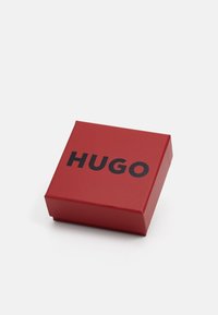 Raudonas kvadratinis dovanų dėžutė su juodu „HUGO“ logotipu centre ant dangčio, padėta ant paprastos baltos fono.