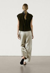 Femme aux longs cheveux blonds en queue de cheval, portant un haut sans manches noir, un pantalon beige à jambes larges et des chaussures noires à enfiler, marchant à l'écart.