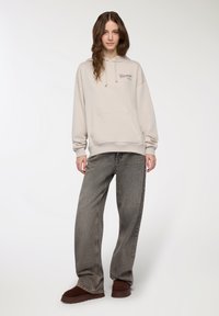 Jasnoszara bluza z kapturem i kieszenią z przodu, zestawiona z ciemnoszarymi spodniami o szerokich nogawkach i brązowymi butami wsuwanymi.