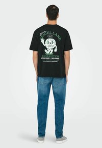 Zwarte katoenen t-shirt met een grafische print op de achterkant, groene tekst en een karakterontwerp. Gecombineerd met blauwe denim jeans.