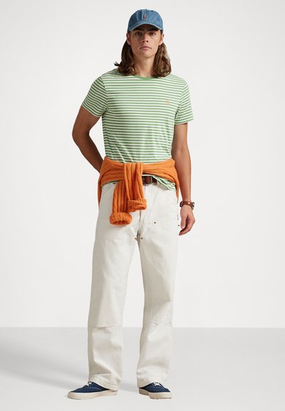 Tricou cu dungi verzi și albe, pulover portocaliu legat în jurul taliei, pantaloni de culoare crem, adidași albaștri și șapcă din denim.