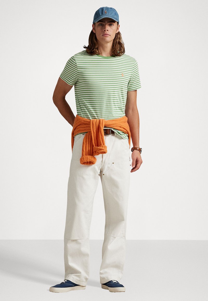 Grün-weiß gestreiftes T-Shirt, orangefarbener Pullover um die Taille gebunden, cremefarbene Hose, blaue Turnschuhe und Jeansmütze.