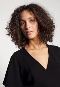Black wrap dress with a v-neck, short sleeves, and textured fabric. Model displays natural curls and minimal makeup against a neutral background.