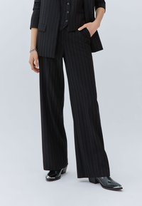 Pantalones de pierna ancha a rayas negras, tela sastrera, sutil brillo, con una cintura alta y corte recto. Combinados con zapatos negros brillantes.