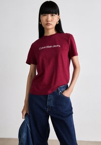 Jeune femme aux cheveux noirs raides portant des boucles d'oreilles en or, un t-shirt bordeaux de Calvin Klein Jeans et un jean bleu ample, tenant une veste en denim.