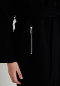 Černý plášť s opaskem, který má na boku stříbrnou kapsu na zip. Tkanina vypadá jemně a má texturu, což zdůrazňuje elegantní design.