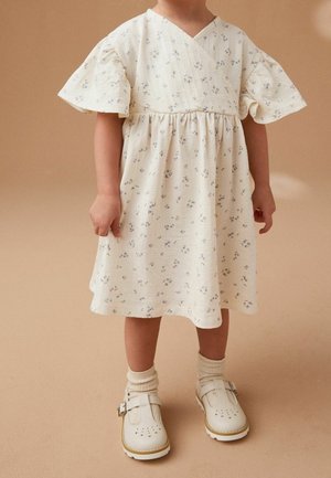 Robe à imprimé floral en crème, dotée de courtes manches volantées et d'une taille plissée. Associée à des chaussures blanches à boucle et des chaussettes crème.