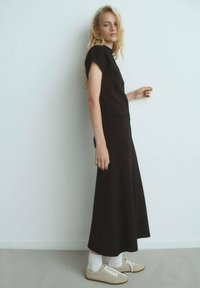 Top negro ajustado con mangas cortas, acompañado de una larga falda negra y fluida. La modelo lleva zapatillas beige con calcetines blancos. Diseño minimalista.