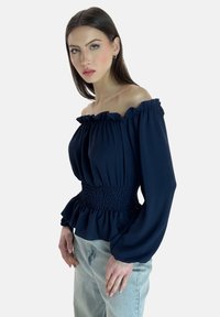 Marineblaue Off-Shoulder-Bluse mit Rüschenbesatz, tailliertem Smock-Einsatz und langen, weiten Ärmeln. Hergestellt aus strukturiertem, weichem Stoff.